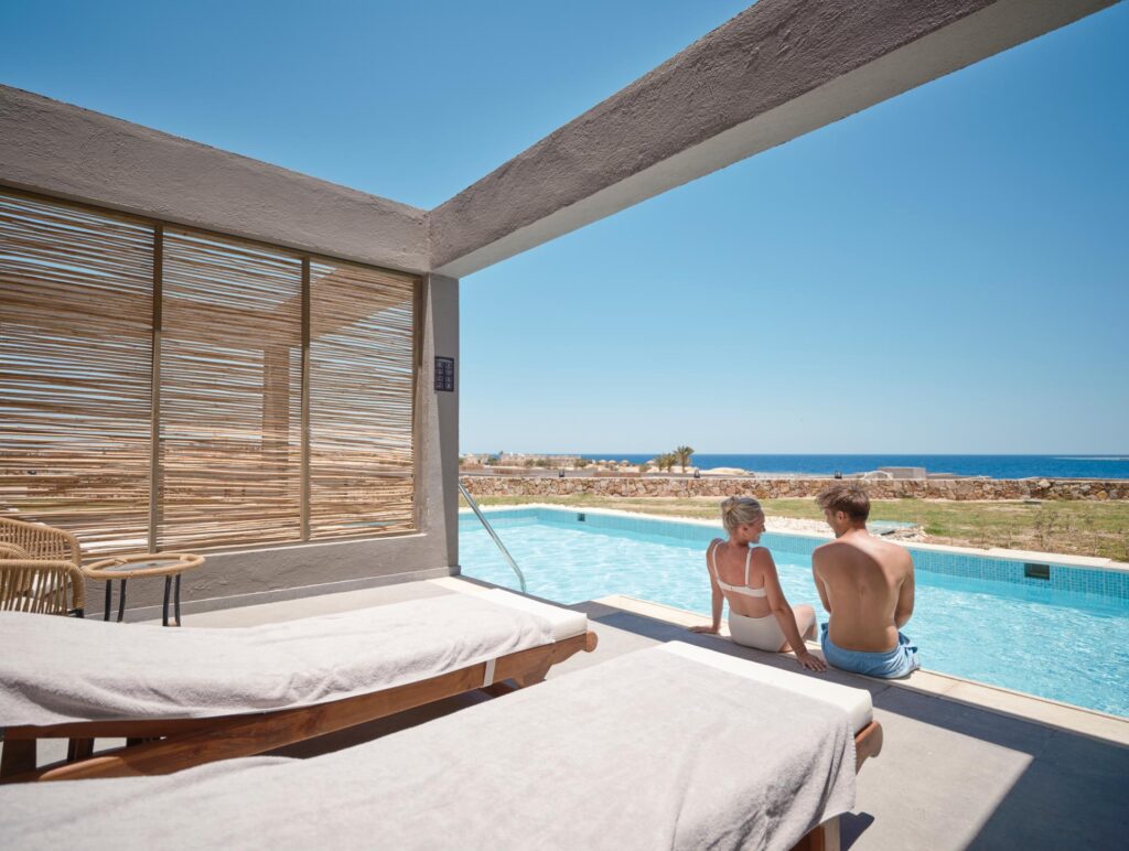 Ein Paar sitzt am Pool mit den Füßen im Wasser und schaut aufs Meer. Swim-up Zimmer im Aldiana Club Naga Bay.
