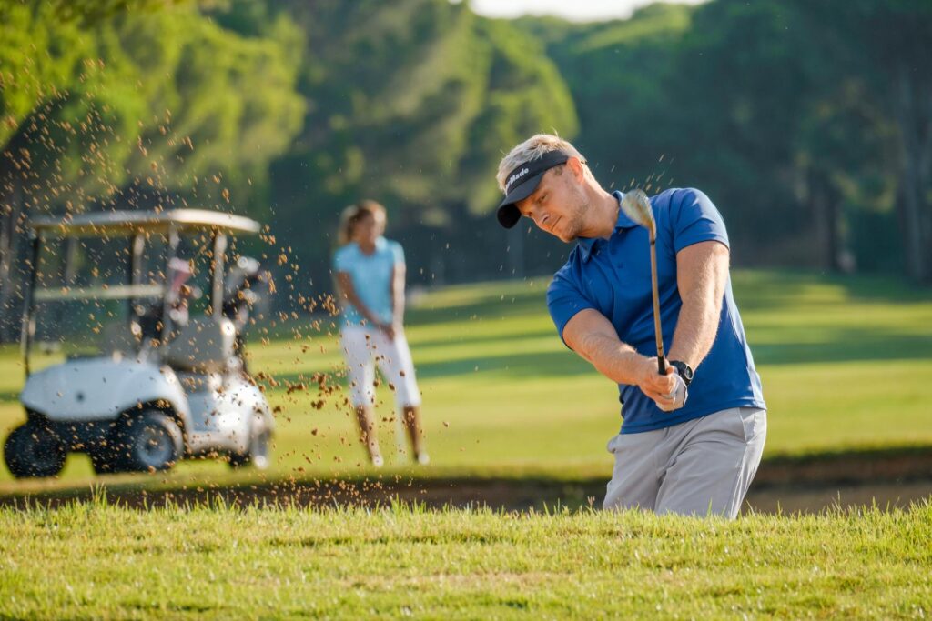Golfspieler schlägt einen Golfball. Im Hintergrund sieht man ein Golfcar und eine weitere Golfspielerin.