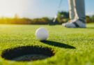 Golfball rollt auf das Loch zu. Im Hintergrund ist der Schläger des Golfspielers zu sehen.