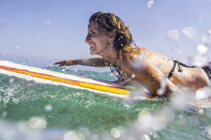 Frau liegt freudig auf dem Surfbrett und schwimmt auf die nächste Welle zur besten Reisezeit auf Fuerteventura zu.