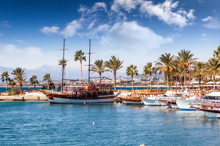 Ein kleiner Hafen in der Türkei mit einigen kleinen und einem größeren Boot auf dem Wasser. Viele Palmen an der Promenade. Blauer Himmel mit nur wenige Wolken im Hintergrund