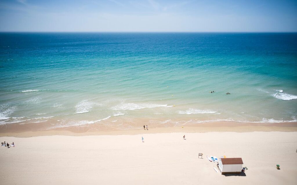 das Meer Andalusiens mit einem Teil des Strandes, einige Menschen im Wasser und am Strand verteilt. Vorne rechts ist ein kleines Häuschen zu erkennen