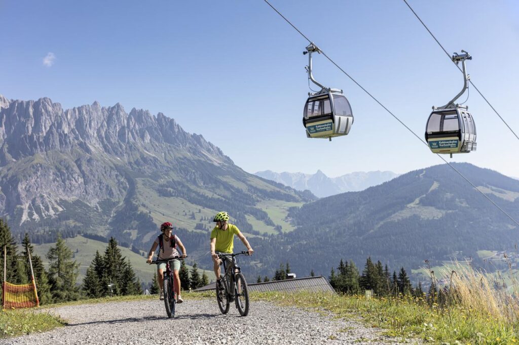 Eine Frau und Mann fahren auf dem Fahrrad im Frühling unter einer Bergbahn in den Alpen durch. 