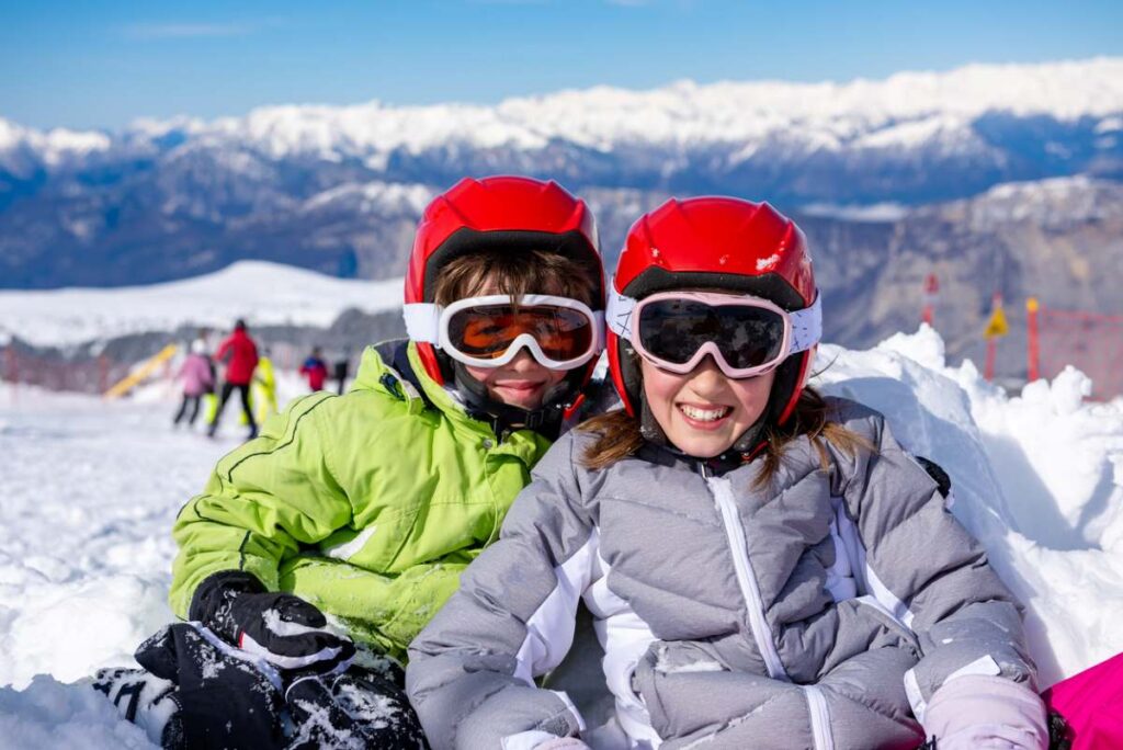 Zwei Kinder mit roten Helmen und Skibrillen im Schnee, im Hintergrund Berge.
