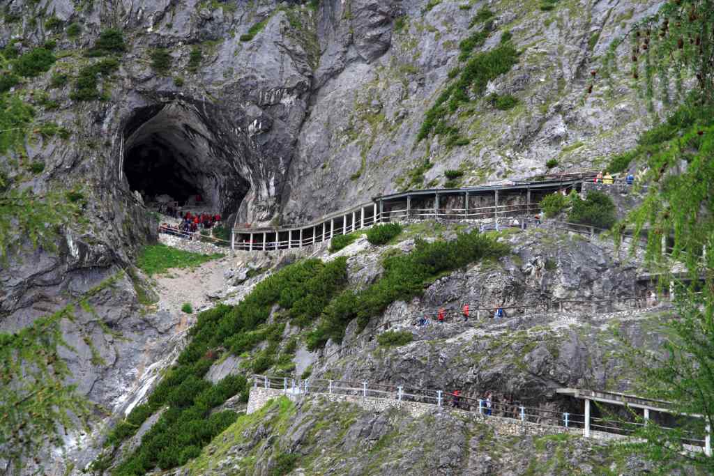 Eingang einer Höhle mit Wanderern auf einem Pfad entlang des Felsens.