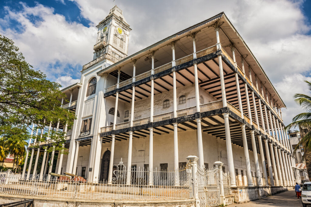 Das House of Wonders in Stonetown, Sansibar bei strahlendem Sonnenschein. Das imposante Gebäude beeindruckt mit seiner kolonialen Architektur und weißen Fassade, umgeben von üppiger grüner Vegetation.