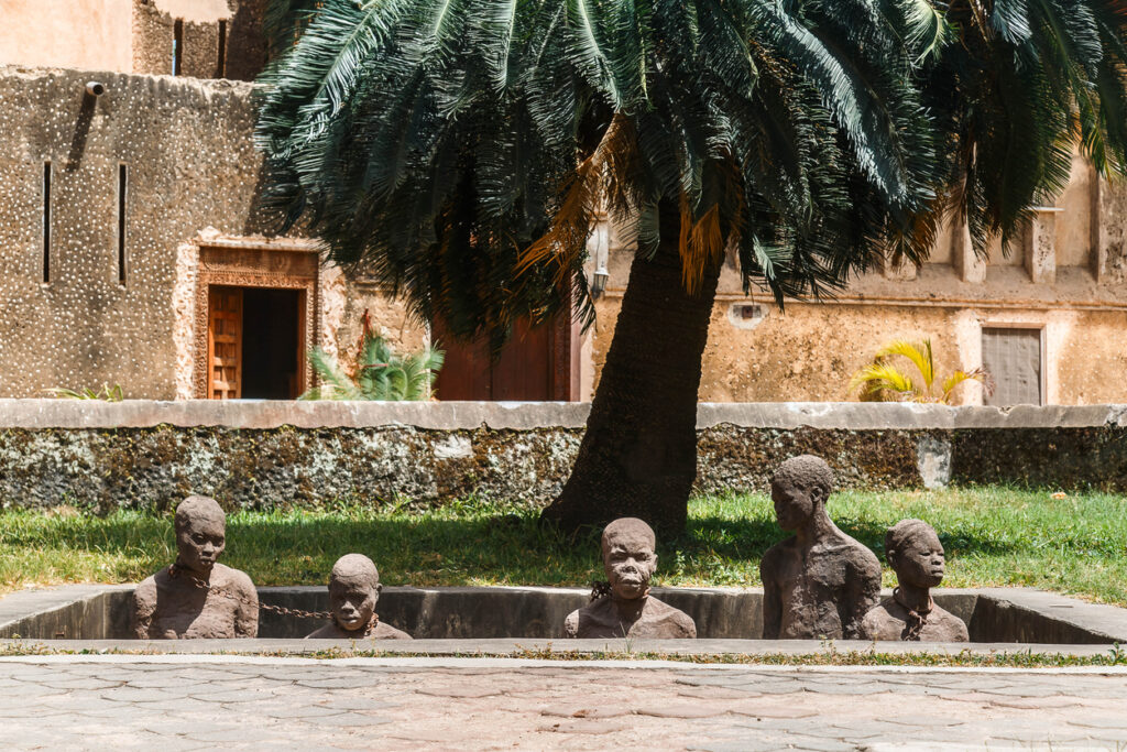 Das Sklaven Denkmal in Stonetown, Sansibar, zeigt mehrere steinerne Figuren, die in einem sandgefüllten, offenen Graben stehen und mit schweren Ketten um den Hals verbunden sind. Das Denkmal erinnert an die traurige Geschichte des Sklavenhandels in der Region und mahnt zum Gedenken. Im Hintergrund sind grüne Pflanzen und eine Steinmauer zu sehen.