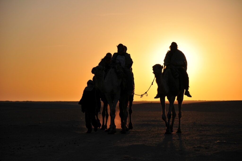 Menschen auf Kamelen in der Wüste bei untergehender Sonne.