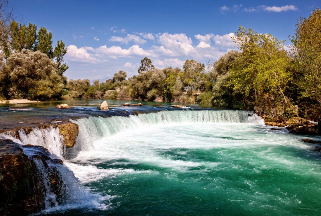 Der brete Wasserfall, umgeben von üppiger Vegetation und fließend türkisfarbenen Wasser unter blauen Himmel mit wenigen Wolken am Horizont.