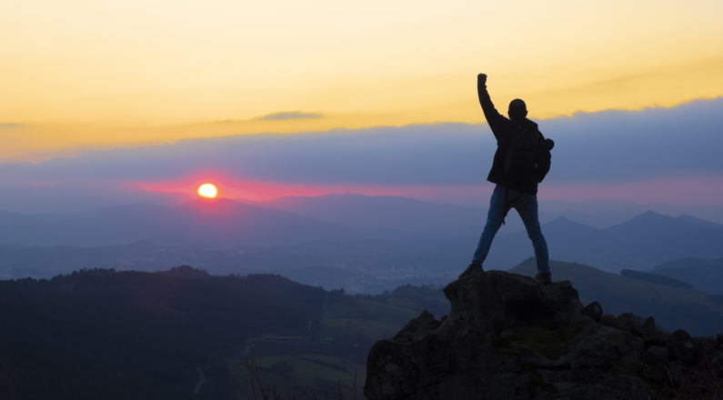 Eine Person steht auf einer Bergspitze und freut sich über den Sonnenaufgang. 