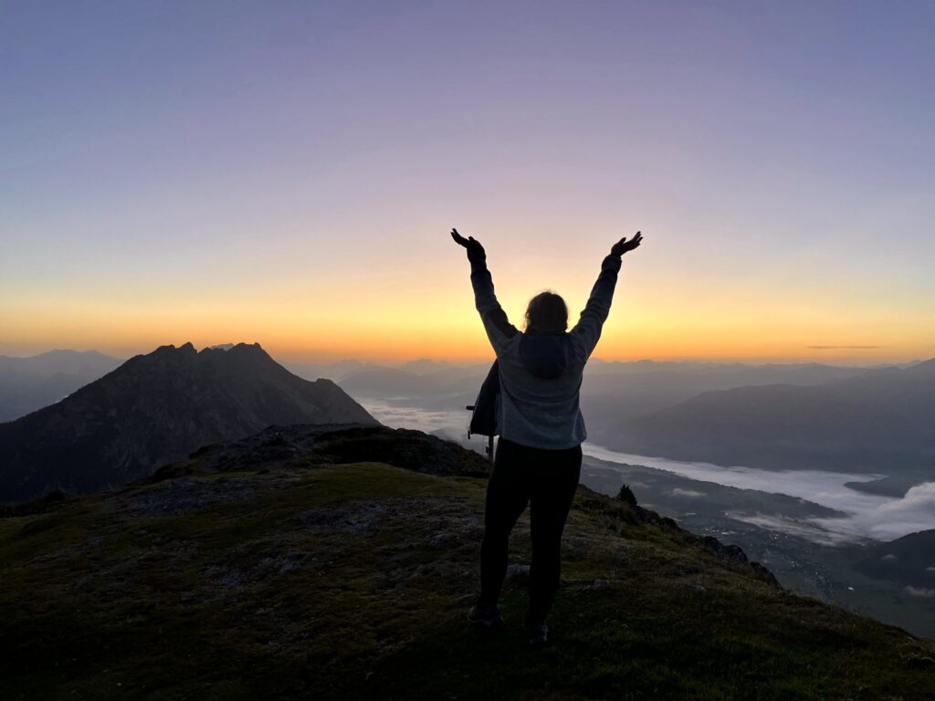 Eine Frau steht auf einer Bergspitze und freut sich über den Sonnenaufgang. 