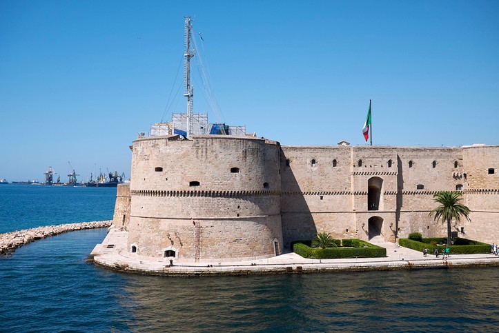 Blick auf die Castello Aragonese, die direkt im Wasser liegt. Gemauertes Gebilde mit wenig grün im Vordergrund und einer italienischen Flagge auf dem Gebäude.