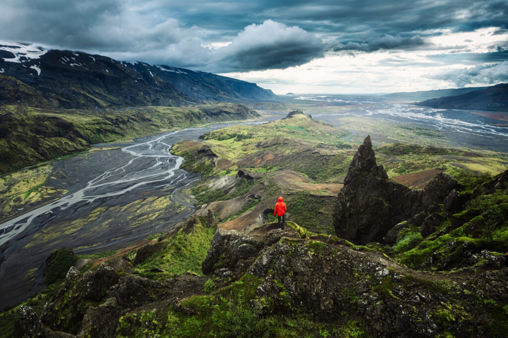 Eine Tpur nach Island lohnt sich in jedem Fall, wenn man Urlaub alleine machen möchte. Hier sieht man einen Wanderer mit roter Jacke auf einem Felsvorsprung steht und ins Tal schaut. 