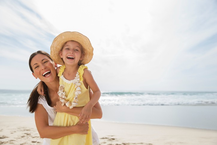 Ein glückliches Mutter-Kind Paar am Strand mit Meer im Hintergrund. Das Kind wird auf dem Arm getragen mit Hut und gelben Kleid sowie einer umgehängten Muschelkette