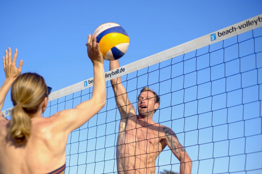 Ein Mann und eine Frau springen direkt am Volleyball-Netz hoch, um beim Beachvolleyball für ihre Mannschaft einen Punkt zu holen. Ein schöner Sport im Urlaub.  