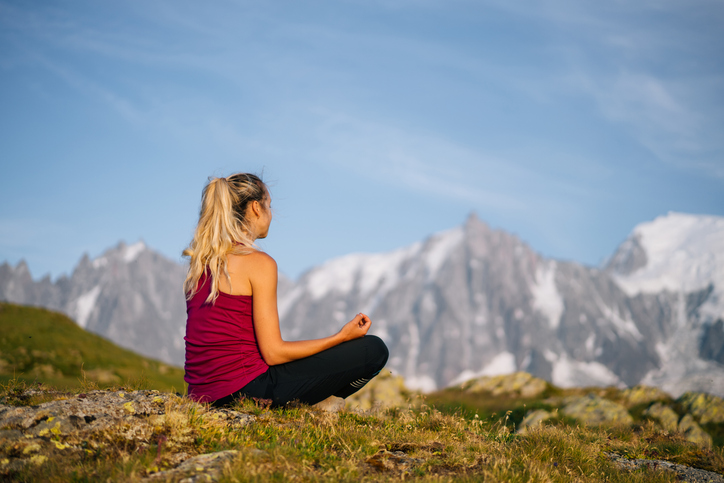 Frau, die auf dem Gras vor einem Bergpanorama sitzt. Yoga und Meditation darf in einem Urlaub mit großem Sportangebot nicht fehlen.